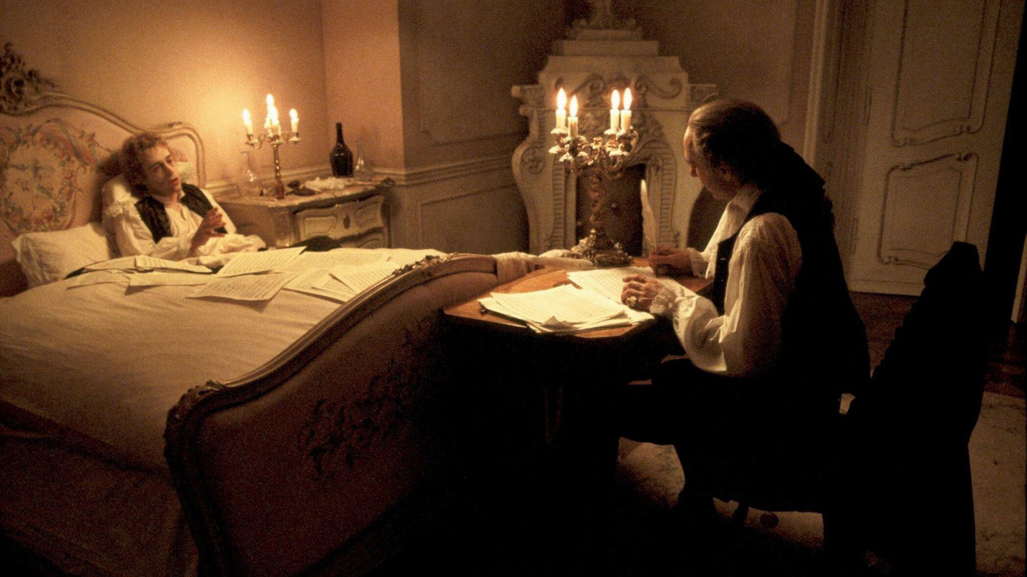 Mozart liegt zugedeckt im Bett, neben dem Bett sitzt Salieri und schreibt Noten in ein Notenblatt